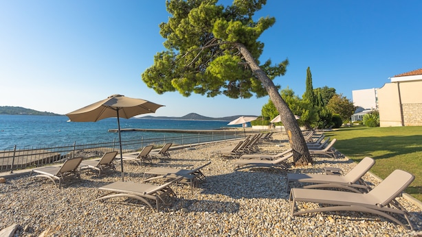 A pebbled beach with lounge chairs and umbrellas, a tree, and a view of the sea and distant hills. - Villa Arausa (Vodice)
