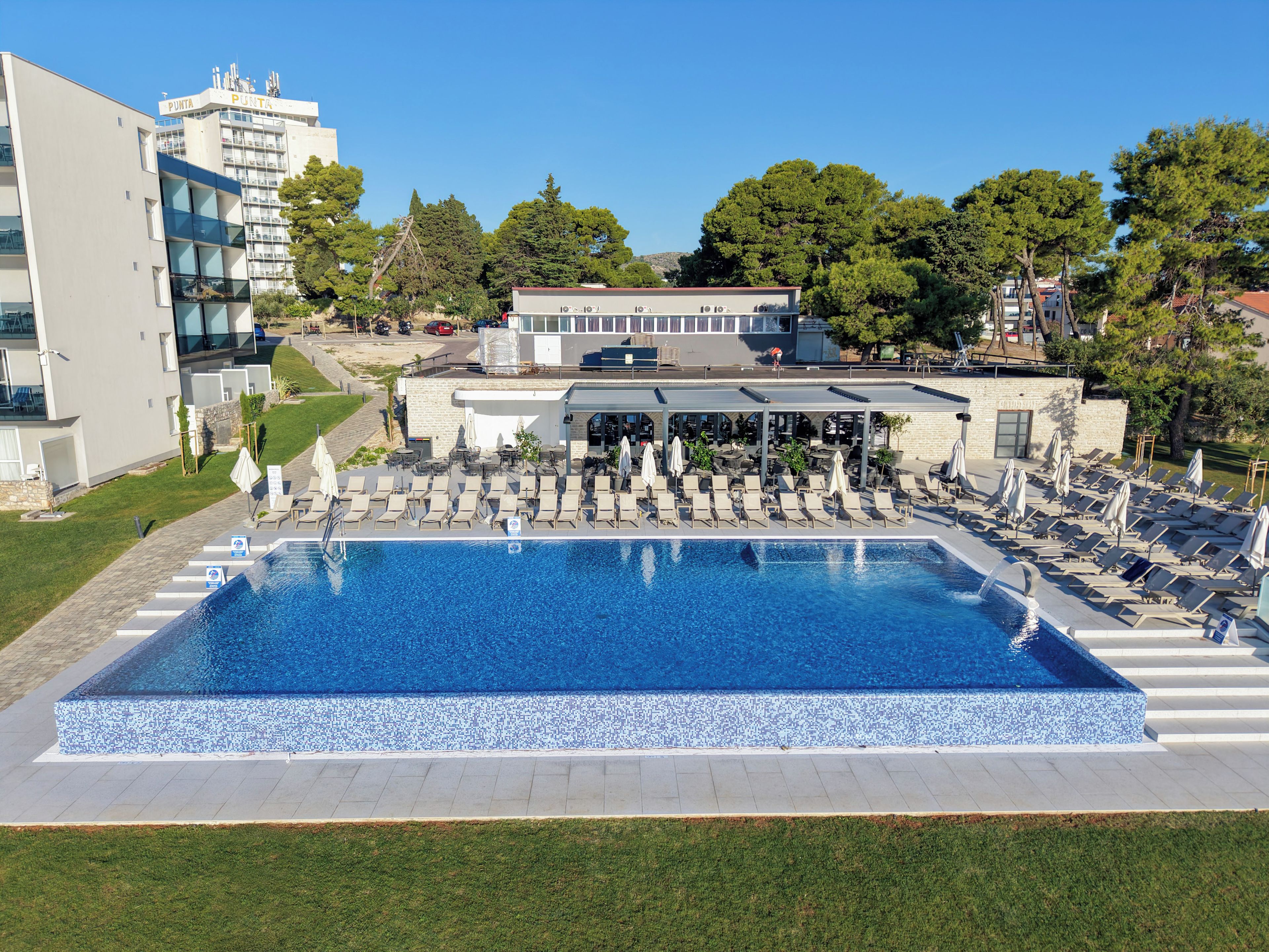 A swimming pool with lounge chairs and umbrellas, surrounded by a building with a covered area and a view of trees and other buildings in the background.