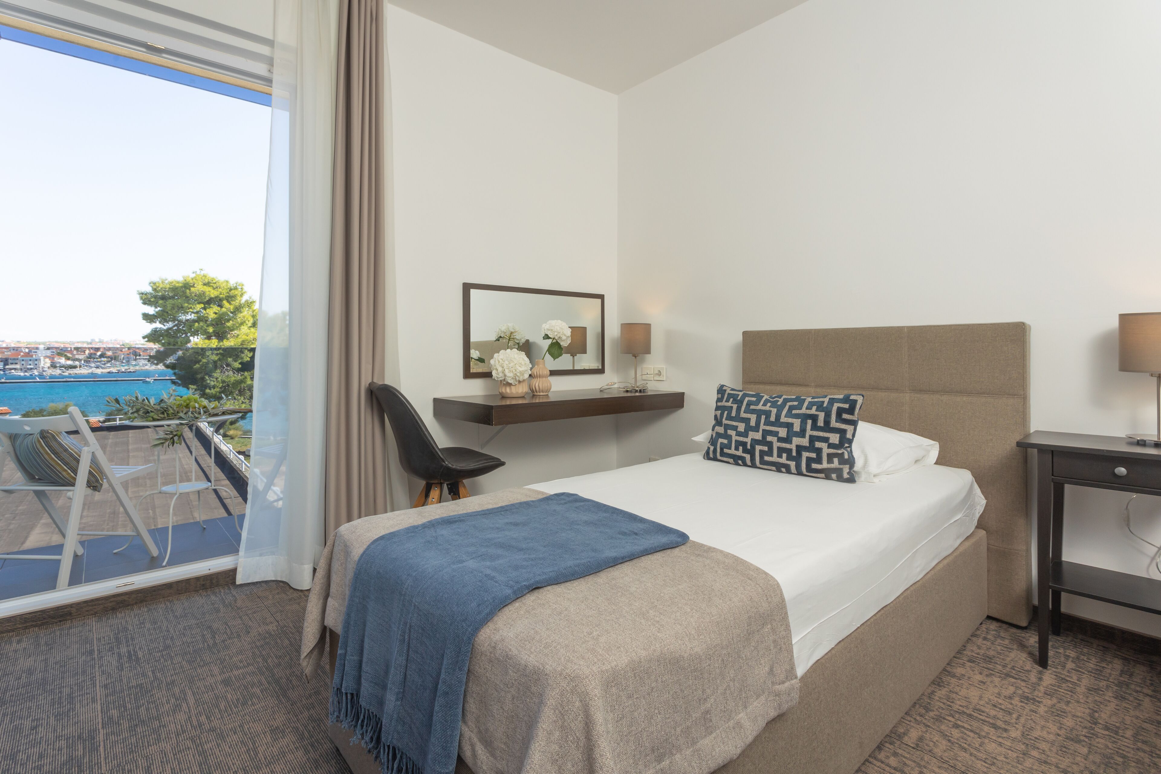 A hotel room with a bed, a desk, a chair, and a view of the sea.