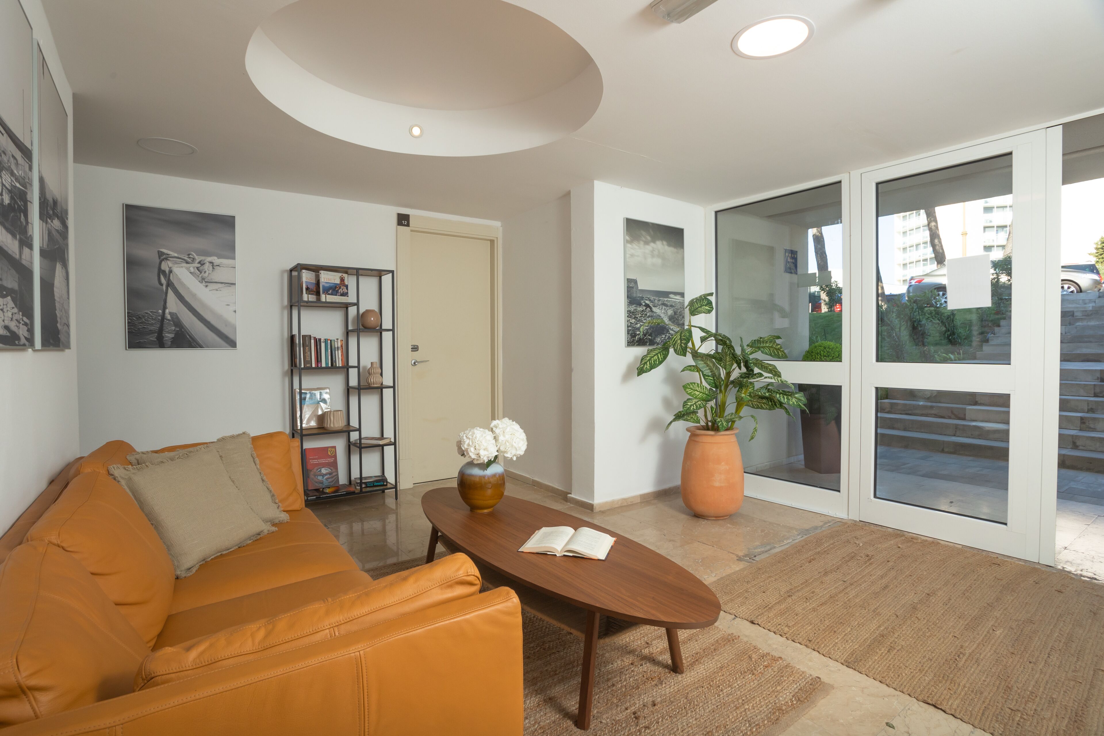 A modern living room with a sofa, a coffee table, a bookshelf, and a large window with a view of the outside.