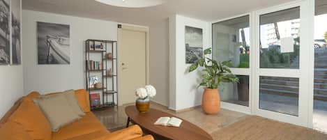A modern living room with a sofa, a coffee table, a bookshelf, and a large window with a view of the outside.