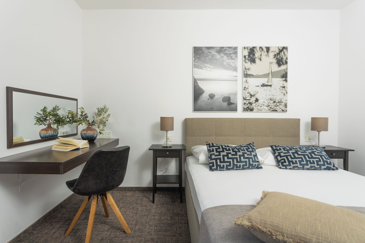 A hotel room with a bed, two nightstands, a desk with a chair, a mirror, and two wall-mounted pictures.