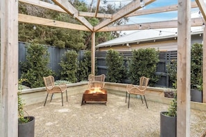 An outdoor patio with a fire pit, three wicker chairs, and a wooden pergola structure.