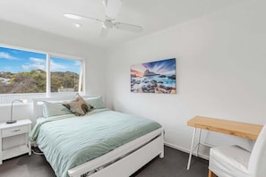 A bedroom with a bed, a desk, a ceiling fan, a window with a view, and a painting on the wall.