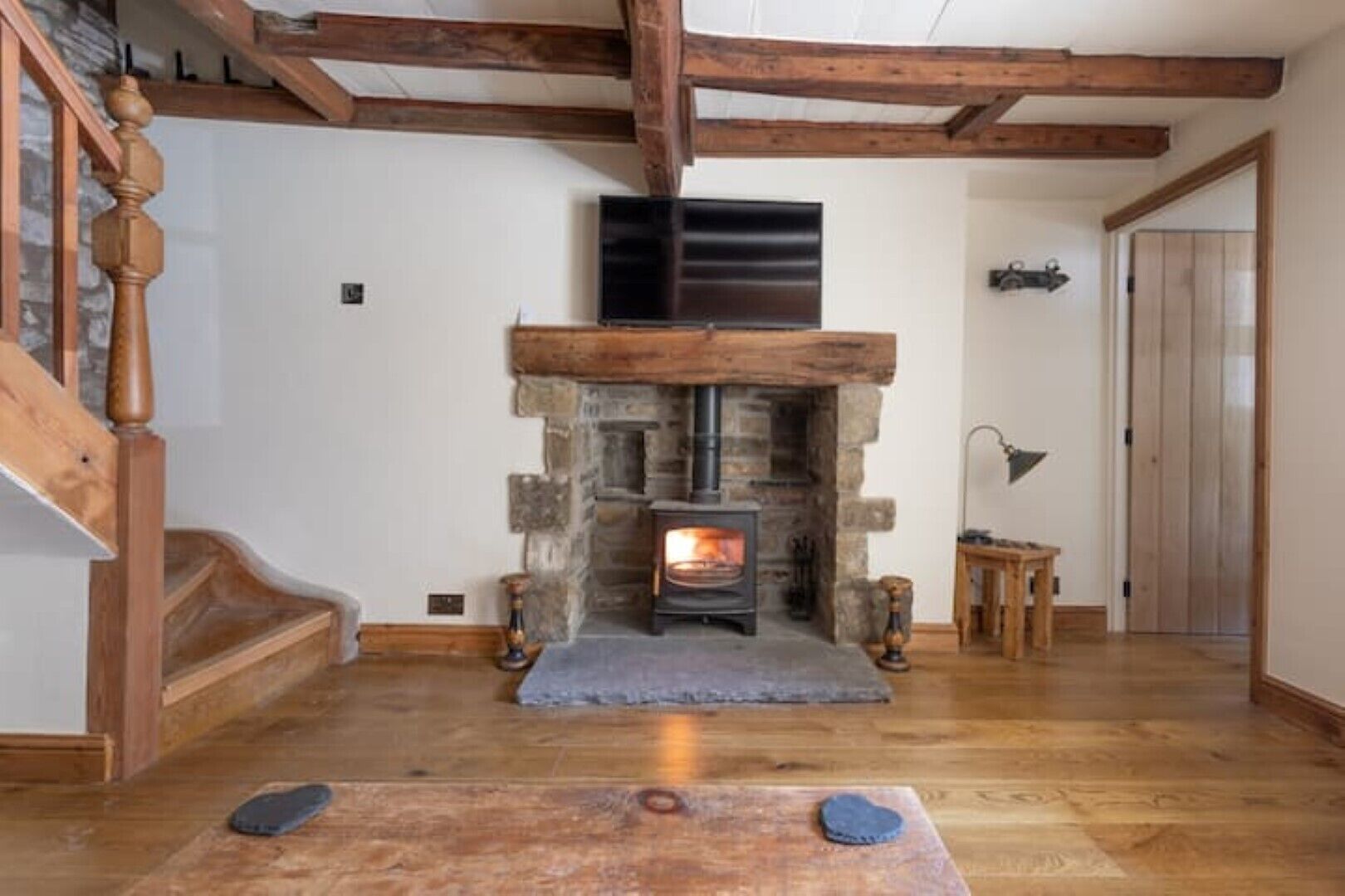 A cozy living room with a stone fireplace, wooden beams, a wooden staircase, and a wooden table with a cushion.