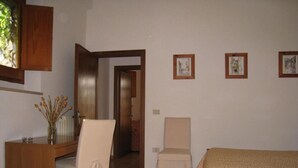 Una camera da letto con un letto, una scrivania, una sedia e un tavolino con un vaso di fiori. Alle pareti ci sono quadri incorniciati e una porta che dà su un'altra stanza.