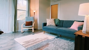 Un salon avec un canapé bleu, un fauteuil, une lampe et une grande fenêtre.