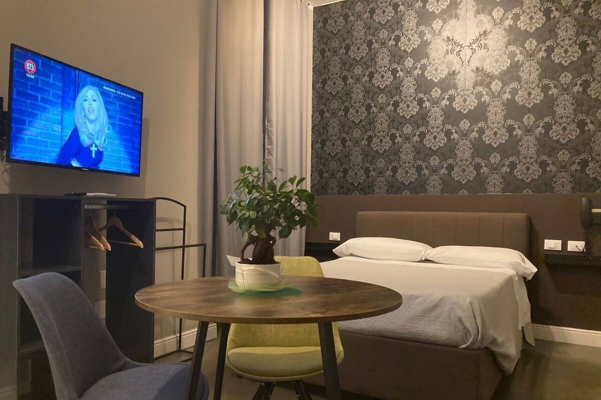 Habitación de hotel con cama, escritorio con computadora, silla, mesa con planta y televisor en la pared.