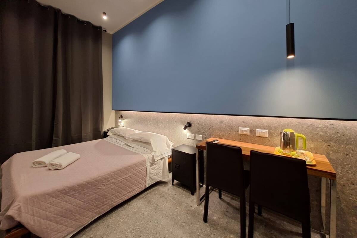 Habitación de hotel con una cama, un escritorio y una silla. Hay una cortina en la ventana y una lámpara sobre el escritorio.