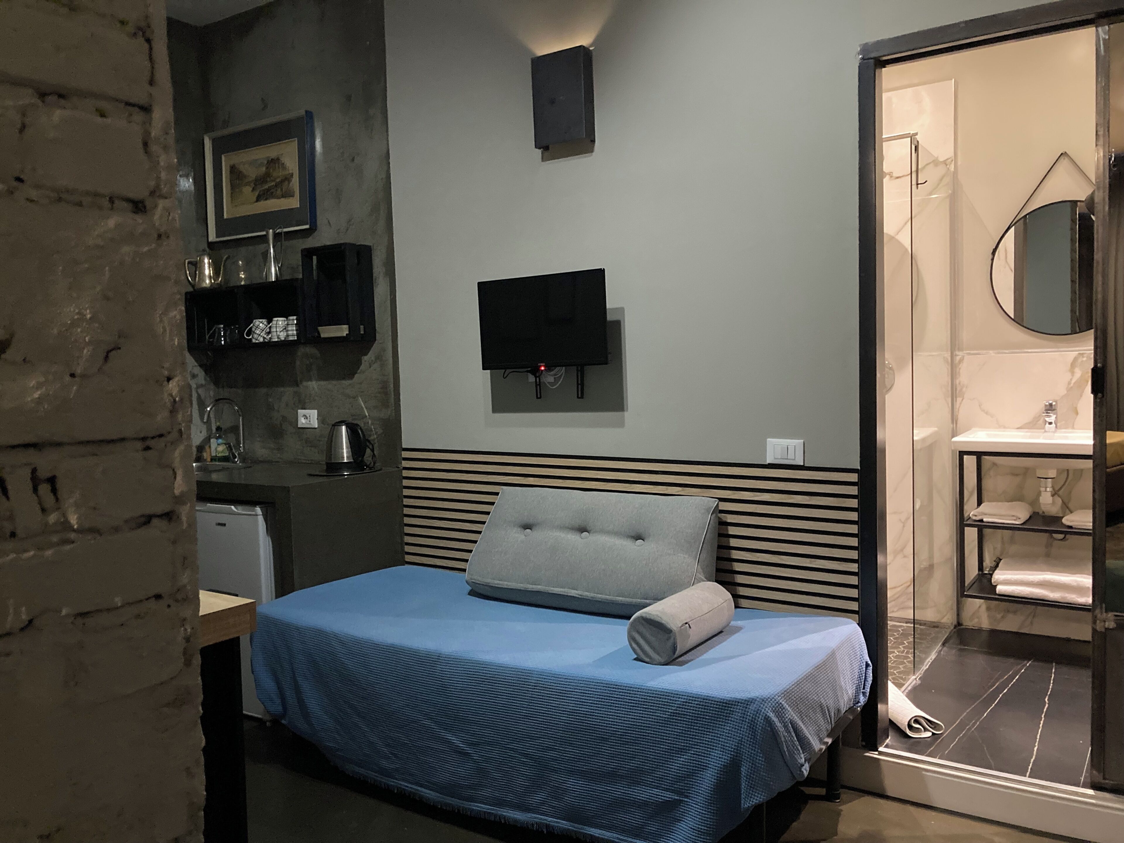 A hotel room with a bed, a television mounted on the wall, a small kitchenette, and a bathroom visible through a glass door.