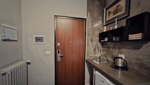 A compact kitchen with a wooden door, a white radiator, a small refrigerator, and a countertop with a kettle and a coffee maker. - Hotel Calypso (Milan)