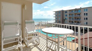 A balcony with a white table and chairs, overlooking a swimming pool and a building with multiple balconies.