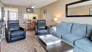 A living room with a sofa, two armchairs, a coffee table with a board game, a dining area with a table and chairs, and a kitchen visible through an open door. - Direct Oceanfront Complex on the No Drive Beach in NSB! (New Smyrna Beach)