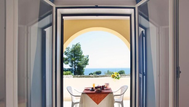 Un umbral abierto que da a un balcón con una mesa dispuesta para dos, con vistas a un paisaje marino y vegetación.