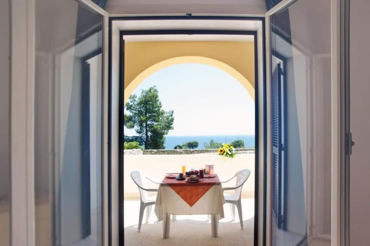 Un umbral abierto que da a un balcón con una mesa dispuesta para dos, con vistas a un paisaje marino y vegetación.