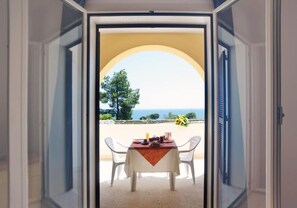 Un umbral abierto que da a un balcón con una mesa dispuesta para dos, con vistas a un paisaje marino y vegetación.