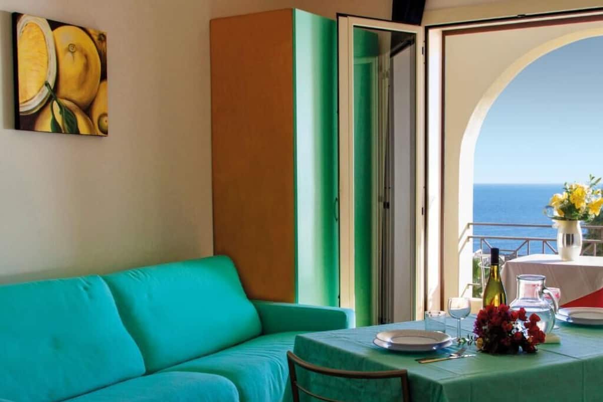 Una habitación con un sofá color turquesa, una mesa de comedor con mantel verde y vistas al mar por una puerta abierta.