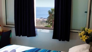 Una habitación de hotel con cama, ventana con cortinas y vistas al mar, además de un florero sobre la mesita de noche.