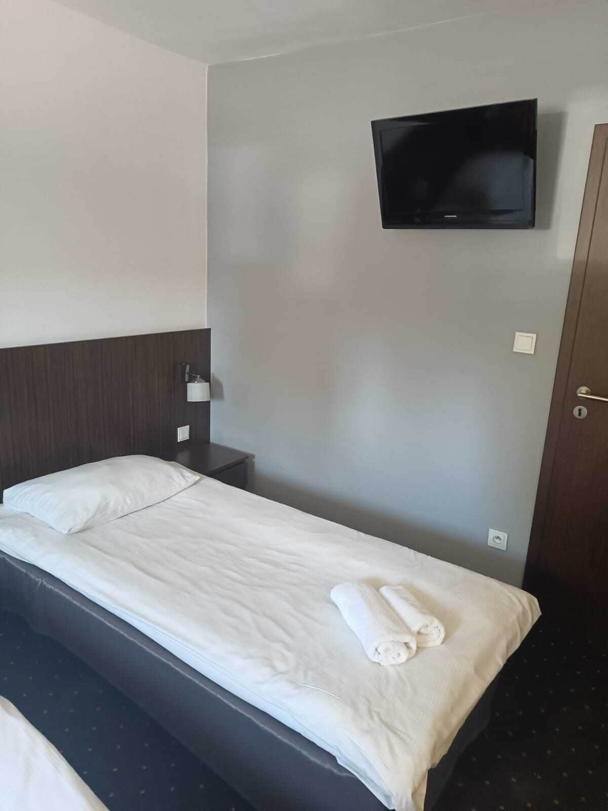 Una habitación de hotel con una cama individual, un televisor de pantalla plana montado en la pared, una mesita de noche y una puerta.