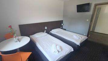 A hotel room with two beds, a small table, a chair, a vase with a flower, and a TV on the wall.