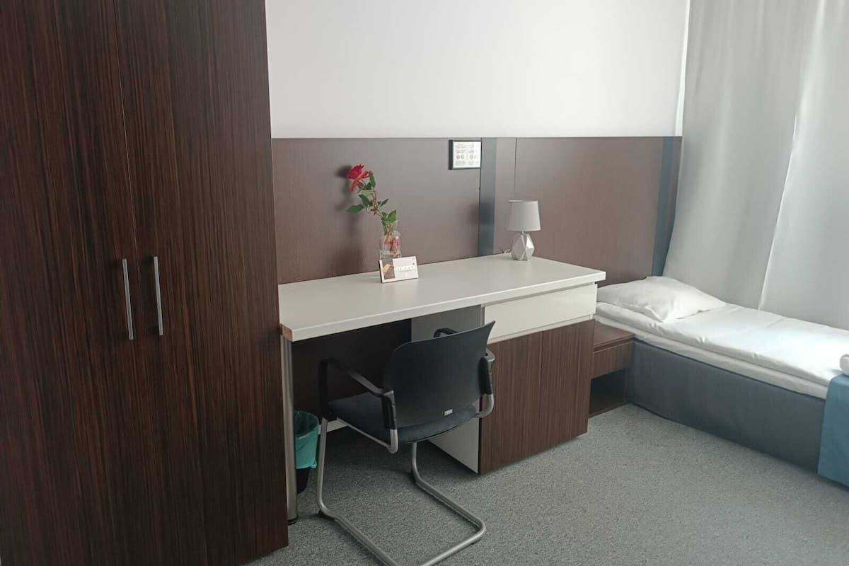 A hotel room with a wooden wardrobe, a desk with a chair, a small bedside table with a lamp, and a single bed.