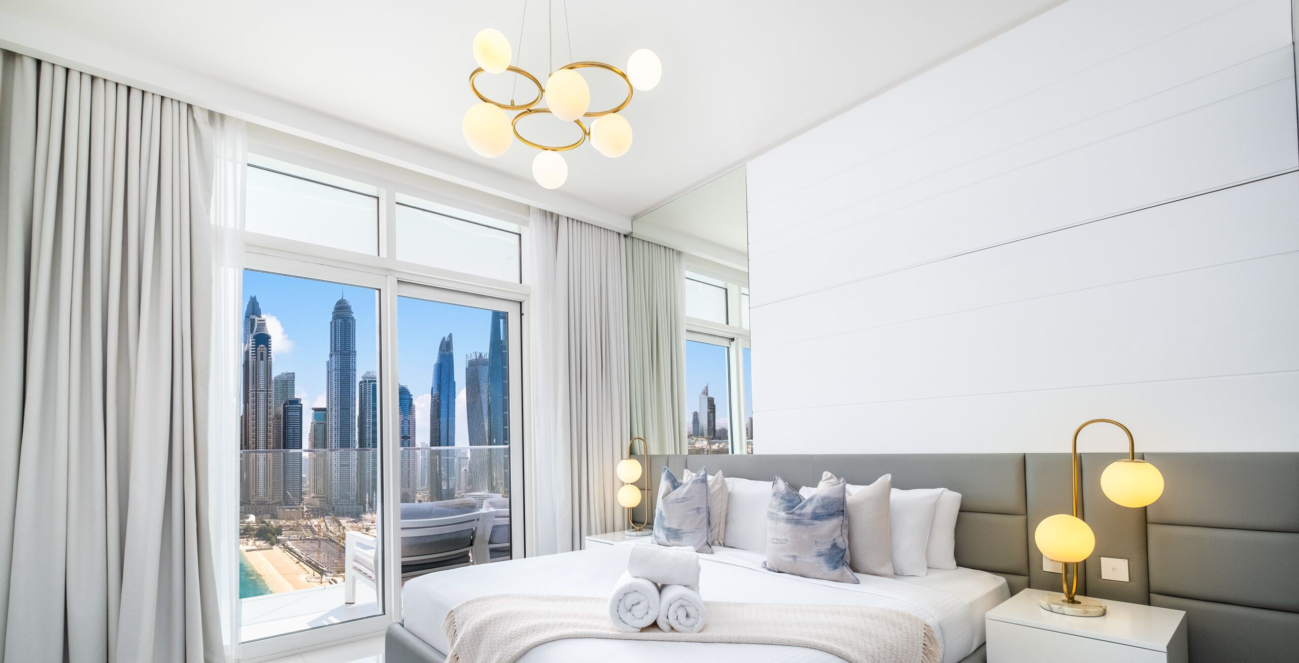 A modern hotel room with a large bed, a bedside table with lamps, and a view of a city skyline through large windows.