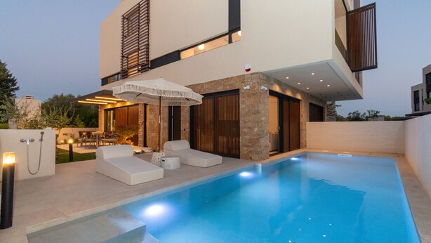 Un área de piscina moderna con una piscina cristalina, sillas de descanso blancas y una zona de estar cubierta con una sombrilla.