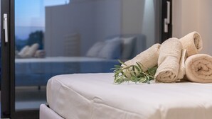 Una cama impecable con toallas enrolladas y una ramita de verdor, una puerta de vidrio que da a un balcón con vistas a un edificio, y un diseño de habitación moderno.