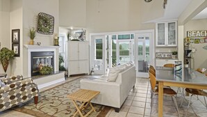 A spacious living room with a fireplace, a white sofa, a dining table with chairs, and a kitchen area with cabinets and a view to the outside.