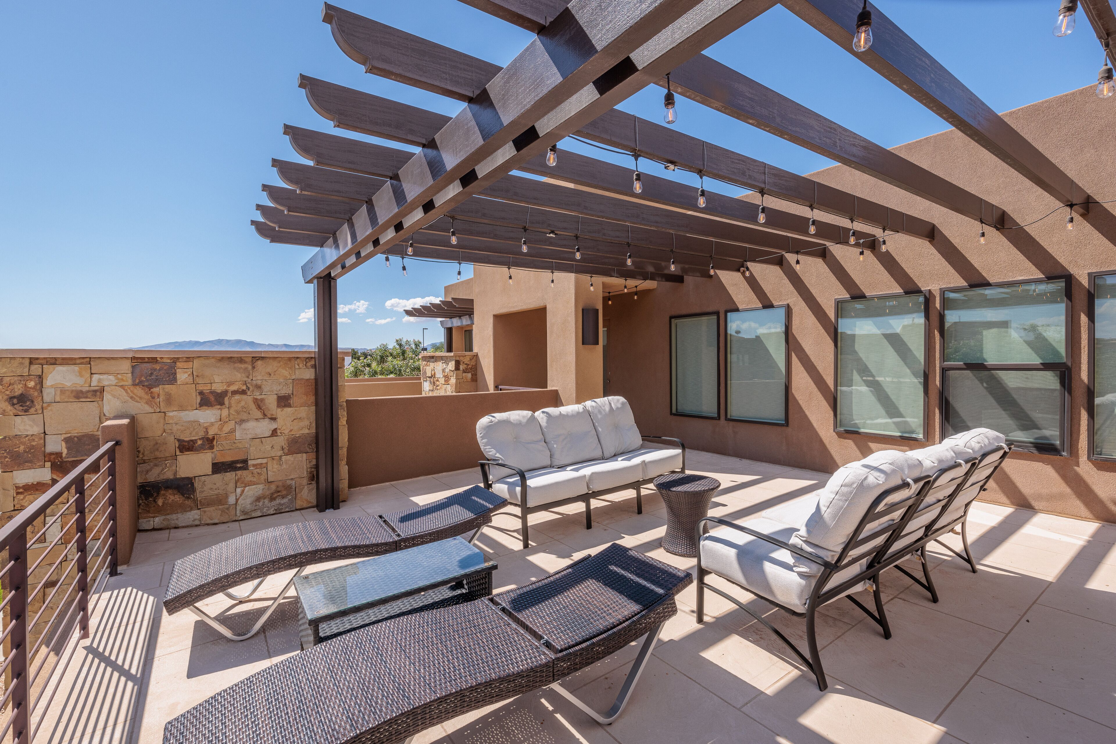 An outdoor patio with wicker furniture, a stone wall, and a pergola with hanging lights.