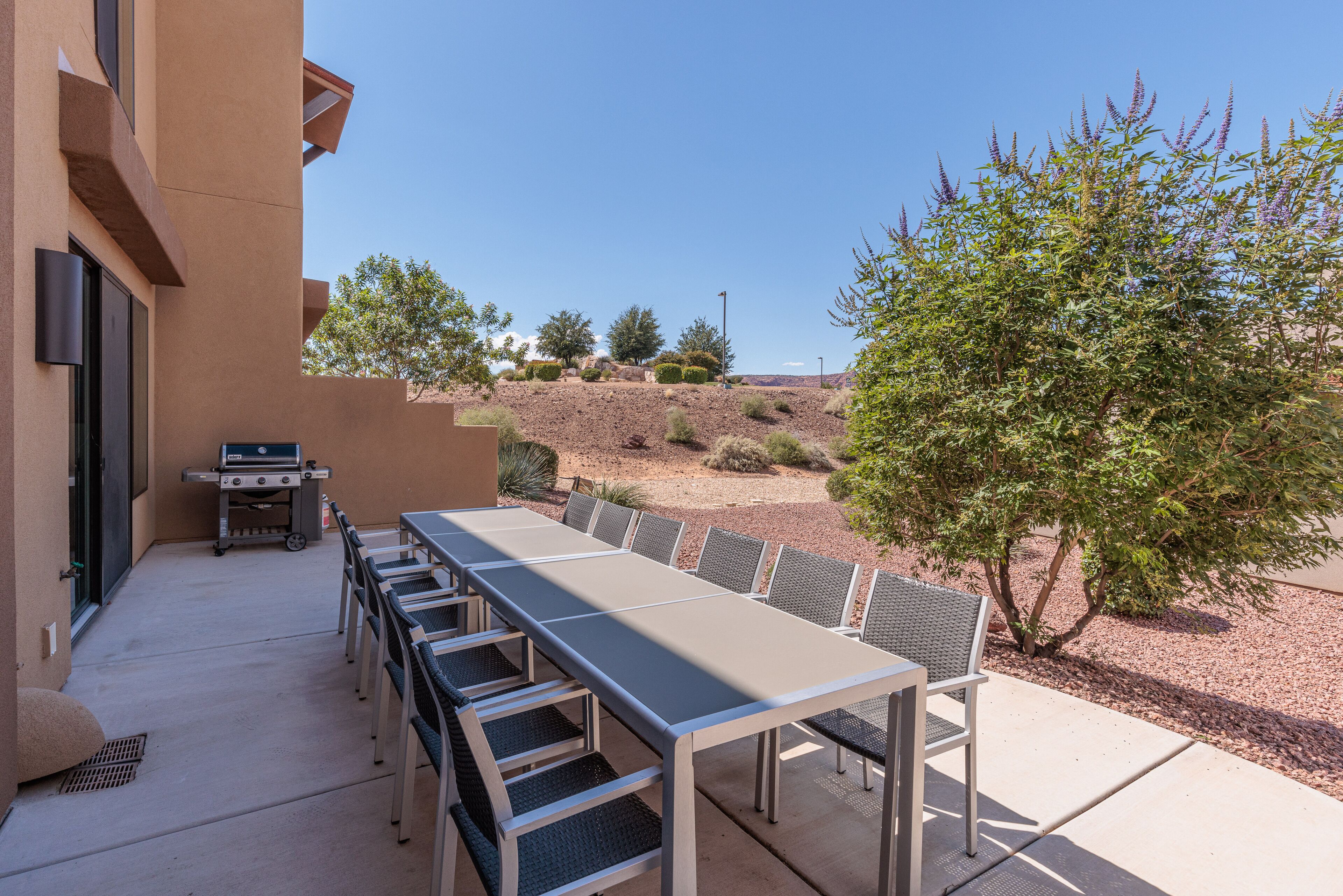 Un patio al aire libre con una mesa larga y sillas, una parrilla y vistas a un área ajardinada con árboles y una colina.