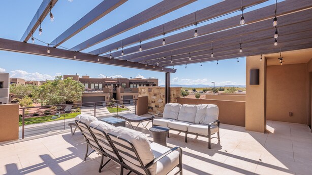 Un patio exterior techado con mobiliario, una pérgola con luces colgantes y vistas de edificios y paisajes circundantes.