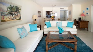 A living room with two white sofas, a glass coffee table, and a blue rug. There is a painting on the wall and a lamp on a side table.