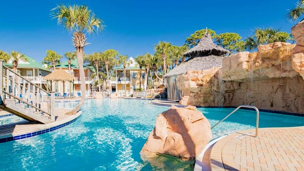 A pool area with clear blue water, palm trees, and a rock formation. - Fully Upgraded Condo, Ideal for Families with Oasis Pool, Tiki Bar & Grill (Pensacola)