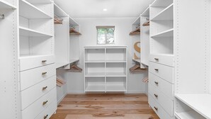 A narrow walk-in closet with white shelving and drawers, a window at the far end, and a ceiling light fixture.