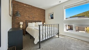A bedroom with a brick wall, a bed, a desk, a lamp, and a window with a view of a hill. - 5 minute walk to PCMR! Stylish condo in the heart of town! Hot Tub, EZ Bus. (Park City)