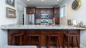 A kitchen with dark wooden cabinets, a stainless steel refrigerator, a microwave, and a granite countertop. There are three wooden stools under the counter. - 5 minute walk to PCMR! Stylish condo in the heart of town! Hot Tub, EZ Bus. (Park City)
