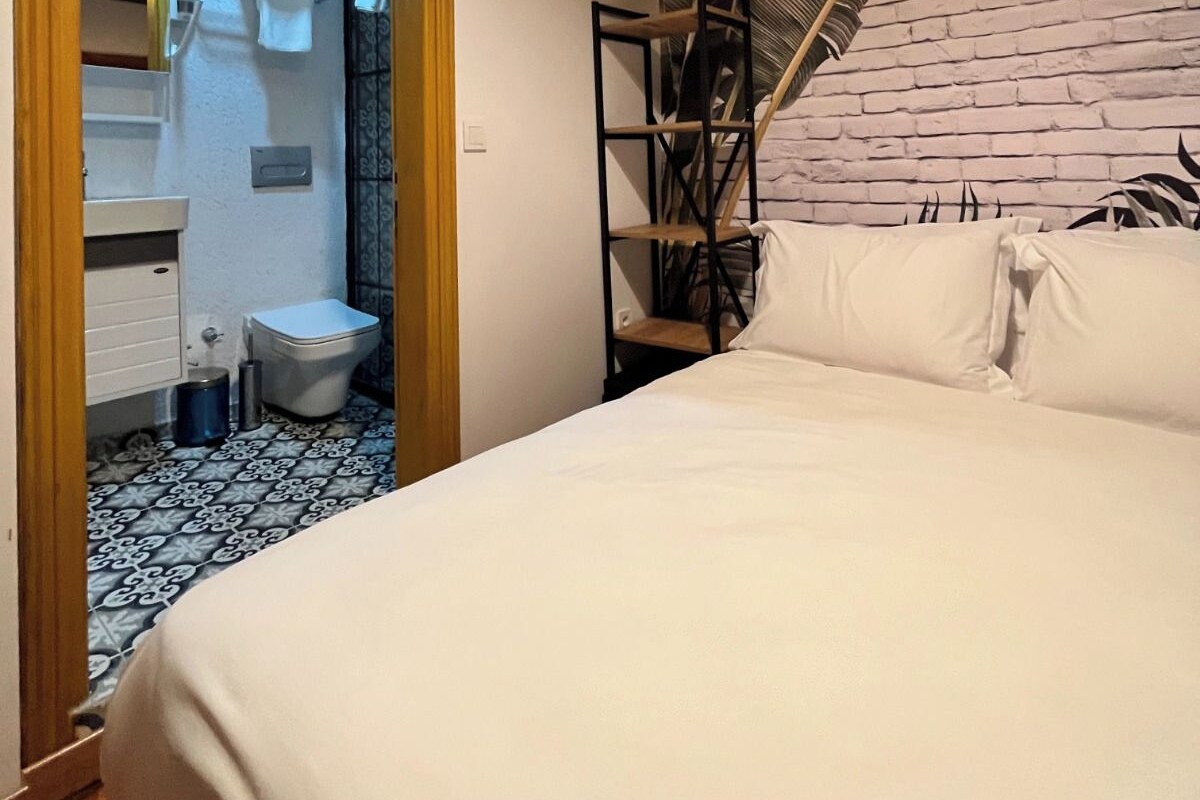Quarto com cama, teto de madeira, parede de tijolos e banheiro visível por uma porta aberta.
