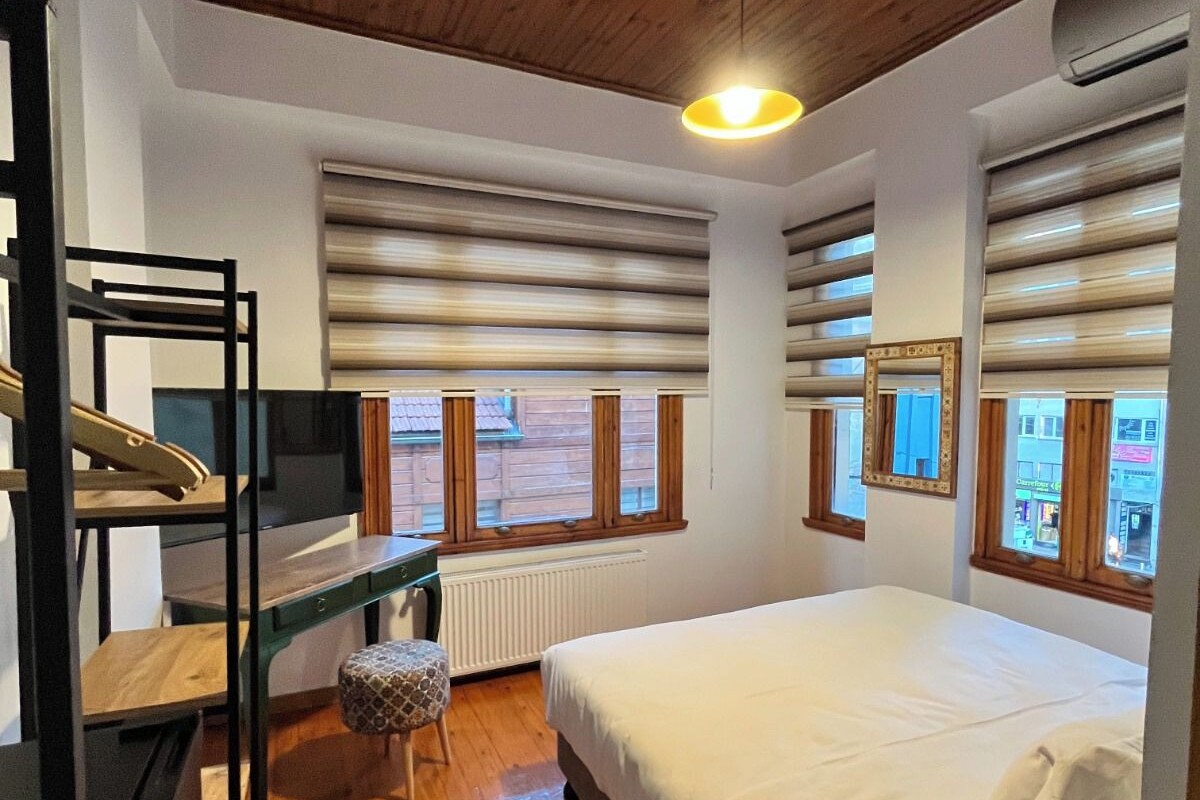 A bedroom with a wooden ceiling, a bed, a desk, and a window with blinds.
