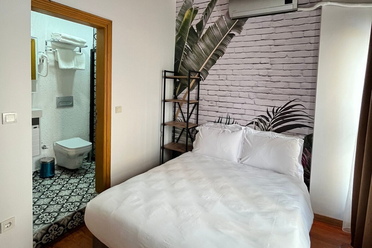 A bedroom with a bed, a wooden ceiling, a brick-patterned wall, a plant, and a bathroom visible through an open door.