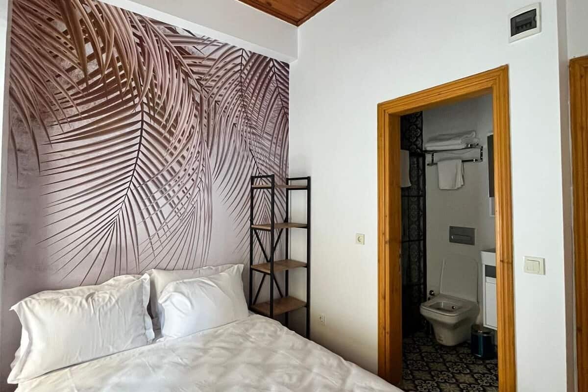 A bedroom with a bed, a wooden ceiling, a wall with a leaf pattern, a shelf, and a door leading to a bathroom.