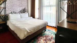 A bedroom with a bed, a desk, a chair, a hanging plant, and a large window with curtains.