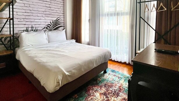 Une chambre à coucher comprenant un lit, un bureau, une chaise, une plante suspendue et une grande fenêtre avec des rideaux.