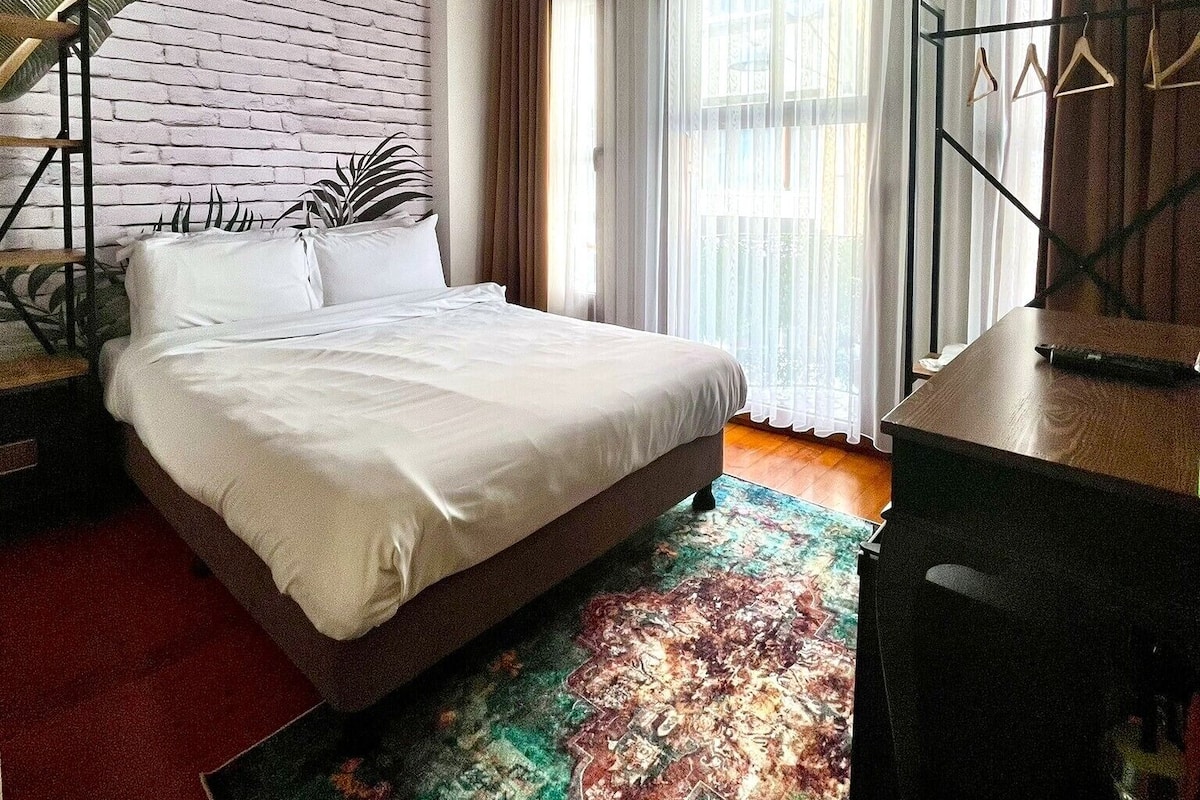 A bedroom with a bed, a desk, a chair, a hanging plant, and a large window with curtains.