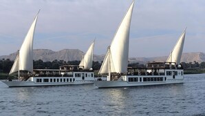 Two large, white riverboats with multiple decks and sails, floating on calm water with a mountainous backdrop. - Dahabeya Farouz El Nil I Nile Cruise (Luxor)