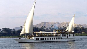 A riverboat with multiple decks and a covered upper deck, sailing on a river with mountains and palm trees in the background. - Dahabeya Farouz El Nil II Nile Cruise (Luxor)
