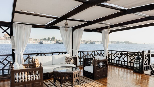 A wooden deck with a sofa and a table, white curtains, and a view of the water. - Dahabeya Farouz El Nil II Nile Cruise (Luxor)