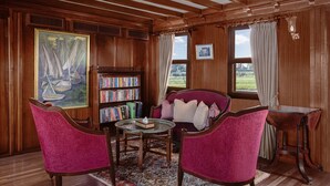 Een kamer met houten lambrisering, een boekenkast, een ronde tafel met stoelen en uitzicht op het landschap door ramen.