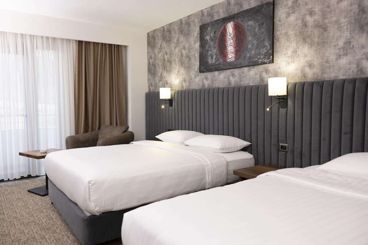 A hotel room with two beds, a grey headboard, a wall-mounted artwork, and a window with sheer curtains.