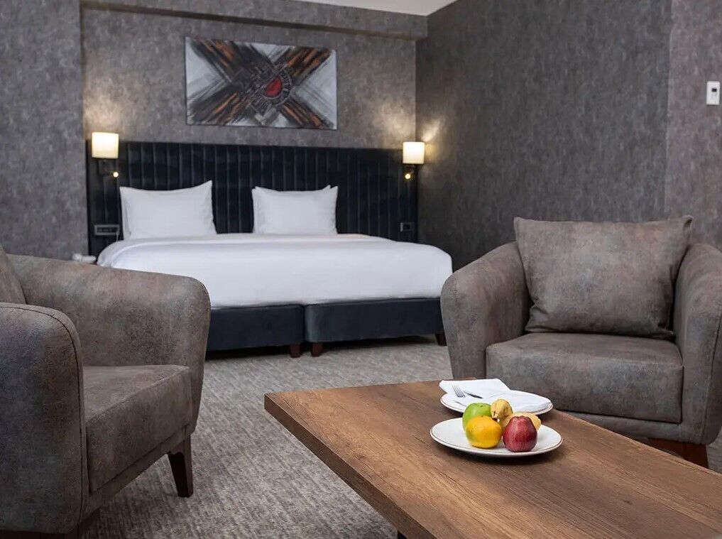 A hotel room with a bed, two armchairs, a wooden coffee table with fruit, and a painting on the wall.